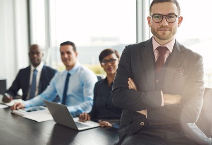 Handsome bearded professional sitting on desk corner beside three coworkers in front of large office window